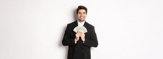 Portrait of happy and pleased handsome man in suit, hugging money and looking satisfied, standing over white background photo