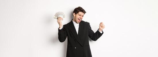 Portrait of handsome and successful businessman in suit, dancing with money, standing against white background photo