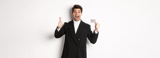 Image of good-looking businessman in black suit, showing thumbs-up and credit card, recommending bank, standing against white background photo