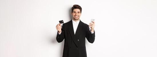 Handsome businessman in black suit smiling, showing credit card and money, standing against white background photo