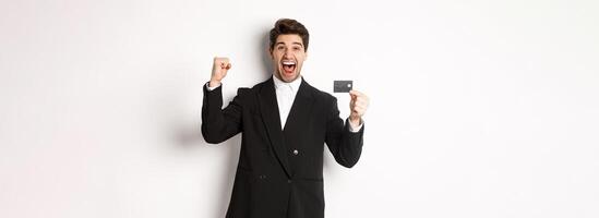Portrait of excited handsome businessman in suit, rejoicing and showing credit card, standing against white background photo