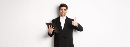 Portrait of confident businessman in black suit, holding clipboard with documents and showing thumb-up in approval, praise good job, standing against white background photo