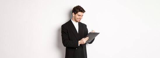 Image of handsome businessman in suit signing documents, looking at clipboard and smiling, standing against white background photo