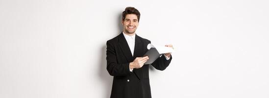 Image of confident businessman in suit looking at documents, holding clipboard and smiling pleased, standing against white background photo