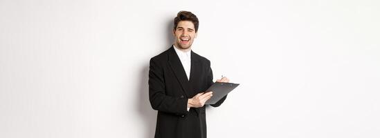 Image of handsome, successful boss in black suit, winking and smiling while signing documents, standing against white background photo