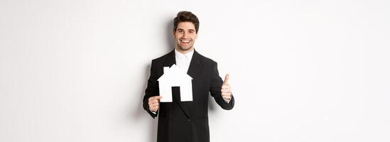 Portrait of confident real estate agent showing house maket and thumb-up, standing against white background photo