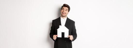 Portrait of handsome man in suit searching for home, holding paper house and looking at upper right corner dreamy, standing over white background photo