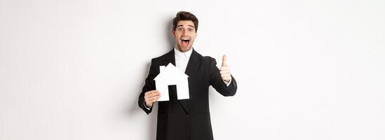Portrait of excited handsome man looking for a home, showing house and thumb-up, recommending real estate agency, standing against white background photo