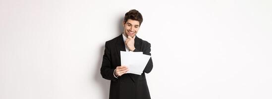 Image of handsome businessman in black suit, looking pleased at documents, reading report and smiling, standing against white background photo