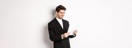 Image of attractive businessman in black suit, reading document and smiling, working on report, standing against white background photo