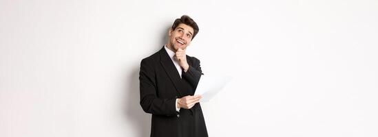 Image of thoughtful, handsome businessman in black suit, holding document and looking at upper right corner, thinking and smiling, standing against white background photo
