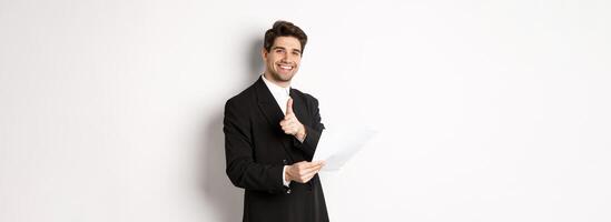 Image of handsome businessman in black suit, holding document and pointing finger at camera, praising good job, standing against white background photo