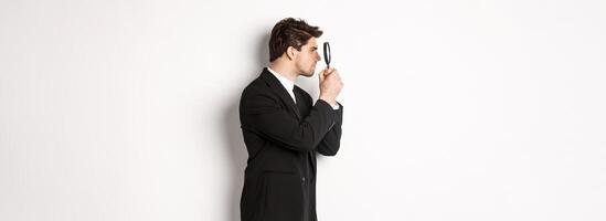 Profile shot of handsome businessman in black suit, looking through magnifying glass and searching for something, standing over white background photo