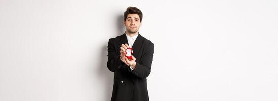 Portrait of handsome man making a proposal, asking to marry him, showing ring, standing in black suit against white background photo