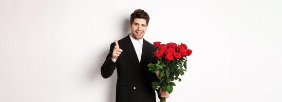 Image of handsome romantic guy in black suit, holding bouquet of roses and pointing at camera, congratulating with holiday, standing against white background photo