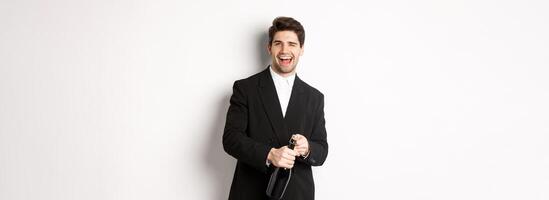 Image of handsome, confident man in black suit, celebrating holiday, open a bottle of champagne and partying, standing joyful against white background photo