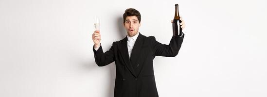 Concept of holidays, party and celebration. Image of handsome man in stylish suit, dancing with bottle of champagne, drinking on new year, standing over white background photo