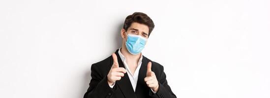 Concept of covid-19, business and social distancing. Close-up of sassy man in trendy suit and medical mask, pointing fingers at camera, standing against white background photo