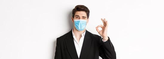 Concept of covid-19, business and social distancing. Close-up of good-looking businessman in trendy suit and medical mask, showing okay sign and winking, standing against white background photo
