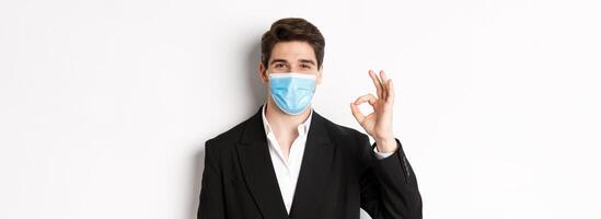 Concept of covid-19, business and social distancing. Close-up of good-looking businessman in trendy suit and medical mask, showing okay sign, recommending something, white background photo