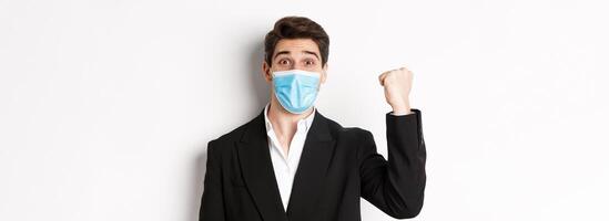 Concept of covid-19, business and social distancing. Close-up of cheerful businessman in medical mask and suit, rejoicing, achieving goal and celebrating, standing over white background photo