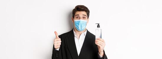 Concept of covid-19, business and social distancing. Close-up of satisfied handsome man in suit and medical mask, showing thumb-up and hand sanitizer, standing against white background photo