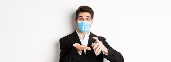 Concept of covid-19, business and social distancing. Image of handsome businessman in trendy suit and medical mask, cleaning hands with hand sanitizer, standing over white background photo
