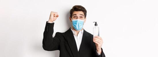 Concept of covid-19, business and social distancing. Close-up of happy man in trendy suit and medical mask, cheering and raising hand up, showing hand sanitizer, white background photo