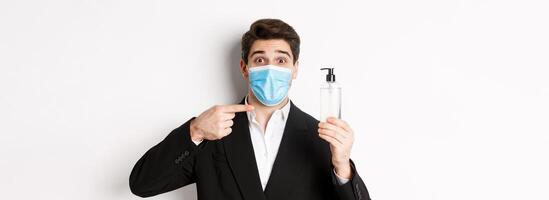 Concept of covid-19, business and social distancing. Close-up of handsome man in trendy suit and medical mask, recommending hand sanitizer, standing against white background photo