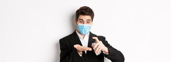 Concept of covid-19, business and social distancing. Image of handsome businessman in trendy suit and medical mask, cleaning hands with hand sanitizer, standing over white background photo
