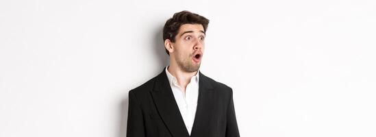 Close-up of impressed guy in trendy suit, open mouth and looking left wondered, standing against white background photo