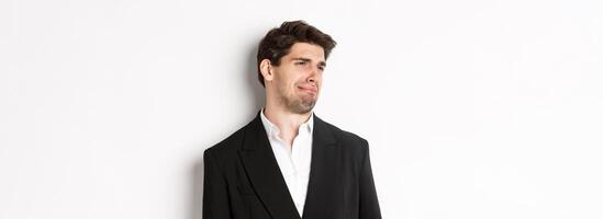 Close-up of disgusted young man in trendy suit, grimacing upset, looking left and standing against white background photo