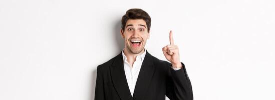 Close-up of handsome businessman in black suit, smiling amazed, showing number one, standing over white background photo