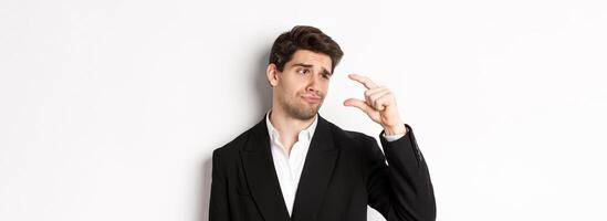 Close-up of handsome businessman in trendy suit, showing something small with disappointment, standing against white background photo