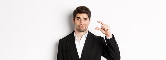Close-up of handsome businessman in trendy suit, showing something small and looking disappointed, standing against white background photo