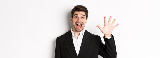 Close-up of handsome businessman in black suit, smiling amazed, showing number five, standing over white background photo