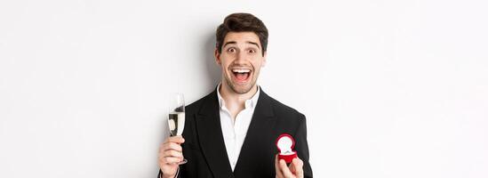 Close-up of handsome man in suit, making a proposal, giving engagement ring and raising glass of champagne, standing against white background photo