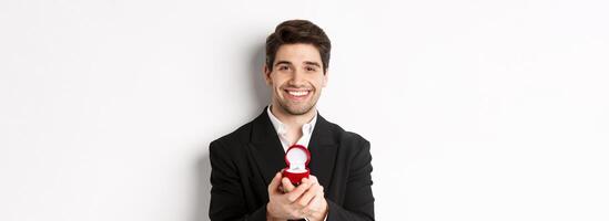 Image of handsome man looking romantic, open small box with engagement ring, making a proposal and smiling, standing against white background photo