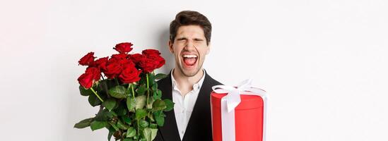 Image of sad guy in suit, got rejected and crying, holding bouquet of roses and present, standing miserable against white background photo