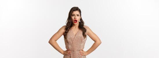 Displeased and shocked woman in party dress and makeup, frowning upset, gasping and looking at something disappointing, complaining, standing over white background photo