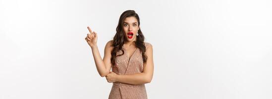 Surprised beautiful woman in luxurious dress and red lips, pointing finger right at promo and starring amazed at camera, standing over white background photo