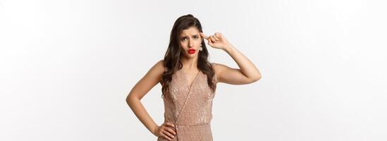 Concept of casino, celebration and party. Woman looking with disdain and roll finger on head, scolding someone, are you stupid gesture, standing in dress over white background photo