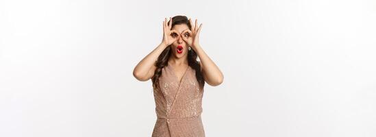 Christmas party and celebration concept. Woman in glamour dress and red lips looking at something amazing, staring through hand binoculars, standing over white background photo