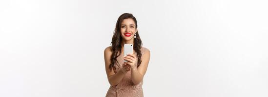 Beautiful young woman in party dress and makeup, taking photo on smartphone, shooting you on phone camera, standing over white background