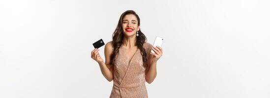 Online shopping and holidays concept. Satisfied and happy woman in elegant dress smiling, using credit card and mobile phone, standing over white background photo