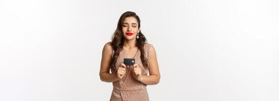 Holidays. christmas party and shopping concept. Satisfied woman in elegant dress rejoicing while showing credit card, smiling happy, standing over white background photo