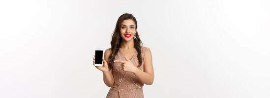 Online shopping. Good-looking woman with red lipstick, luxury dress, pointing finger at mobile screen, showing internet store or app, white background photo