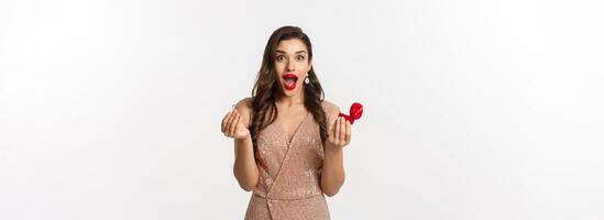 Excited woman holding engagement ring and red box, looking surprised and amazed, receive marriage proposal on date, wearing elegant dress, standing over white background photo