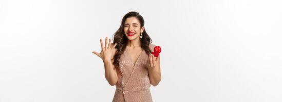 Excited young woman in glamour dress being engaged, showing finger with ring and looking happy, receive marriage proposal, standing over white background photo