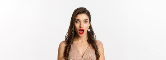 Image of attractive woman with red lips and hairstyle, wearing dress, staring surprised at camera, standing over white background photo